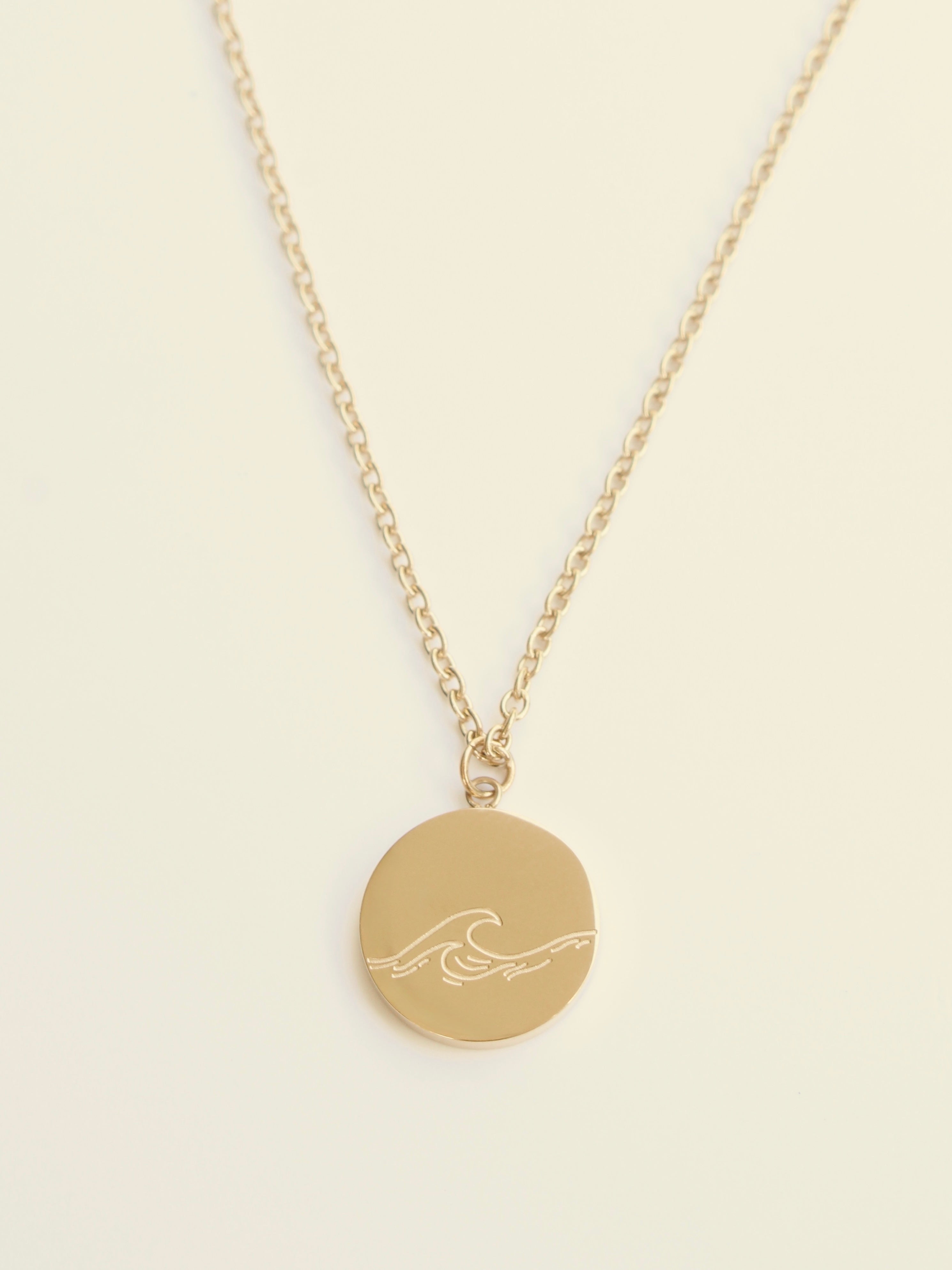 Gold necklace with a round pendant featuring a wave design on a beige background