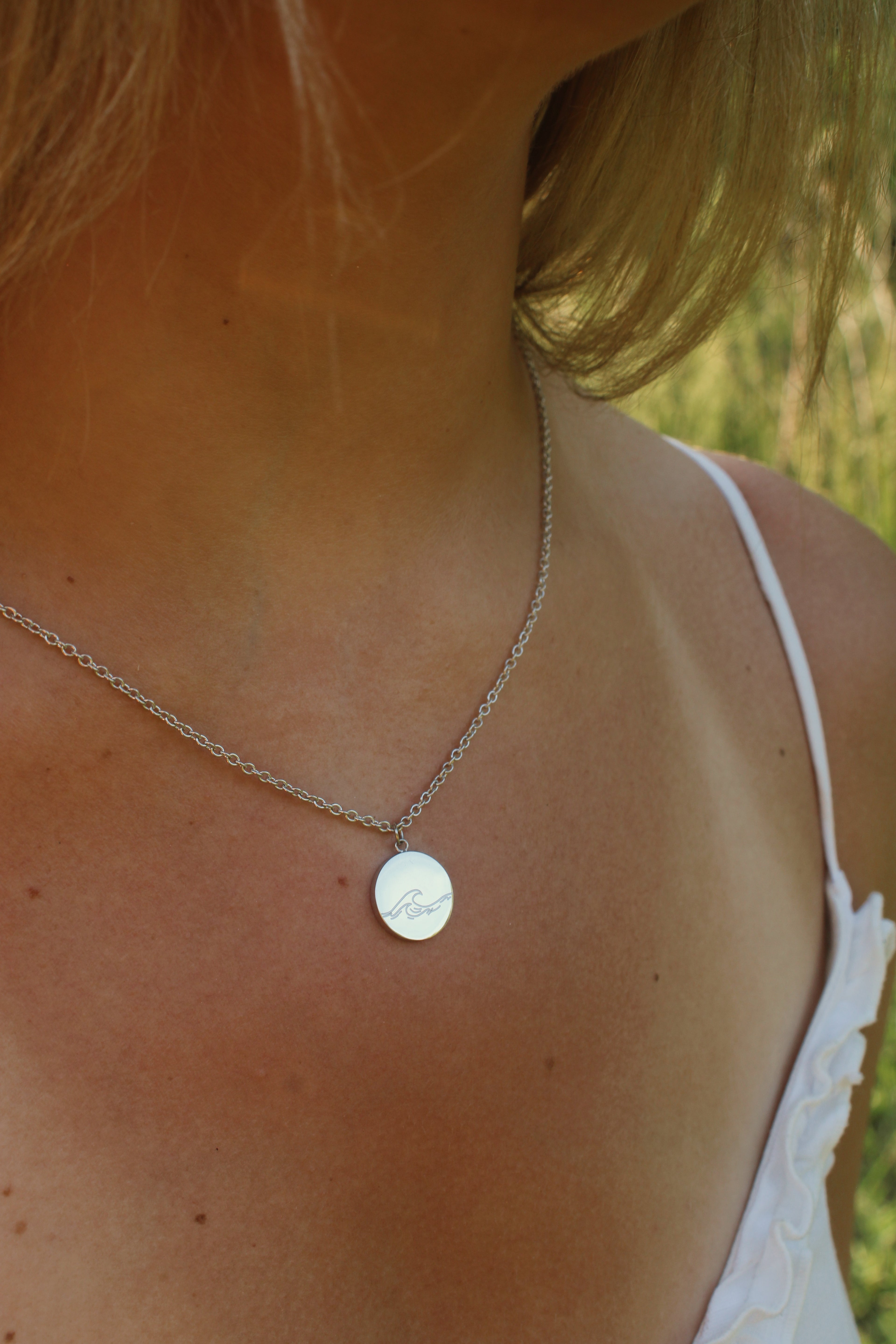 Close-up of a necklace with a round pendant on a person wearing a white top outdoors.